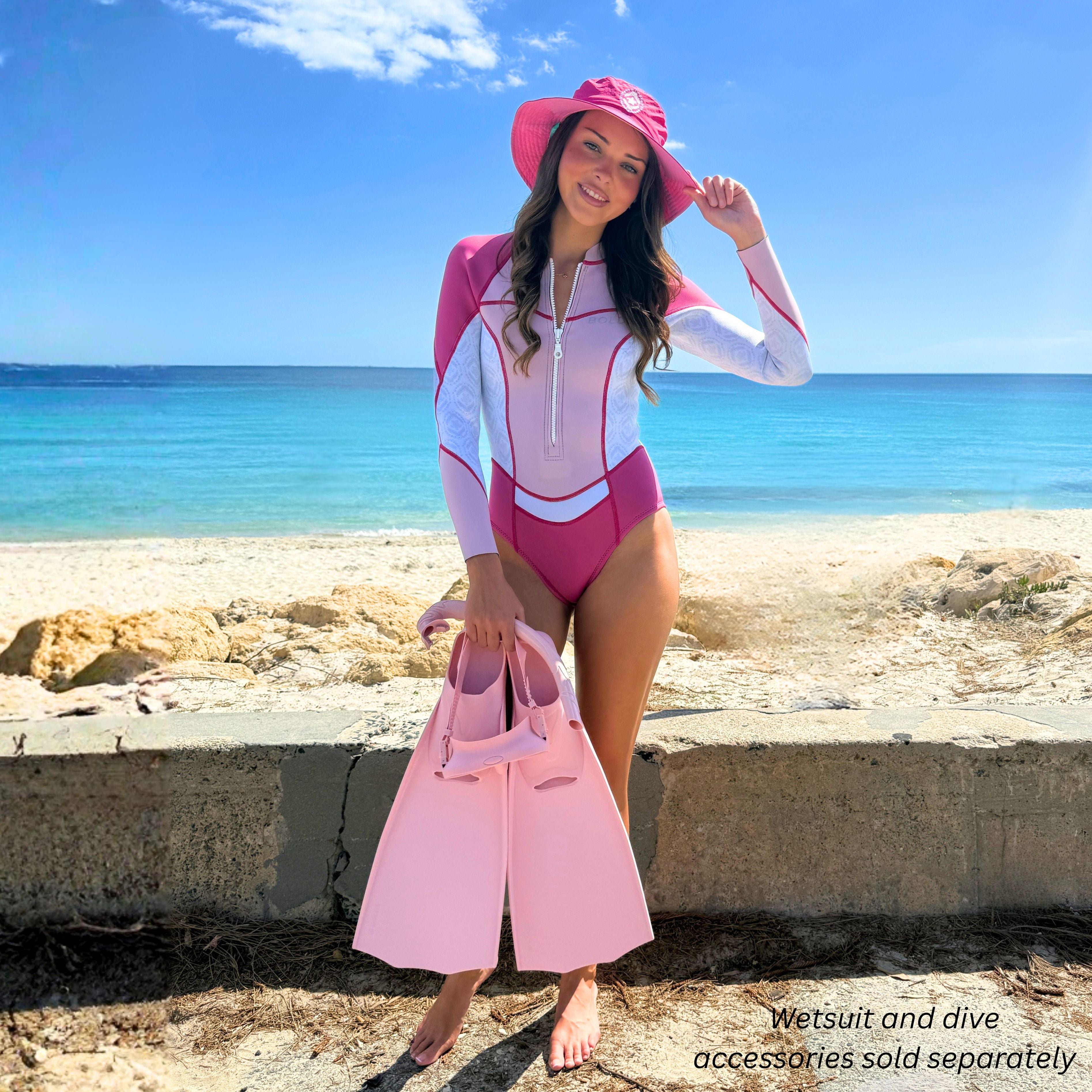 A model in a BOLDE pink and white springsuit with a matching beach hat and dive fins on a beach